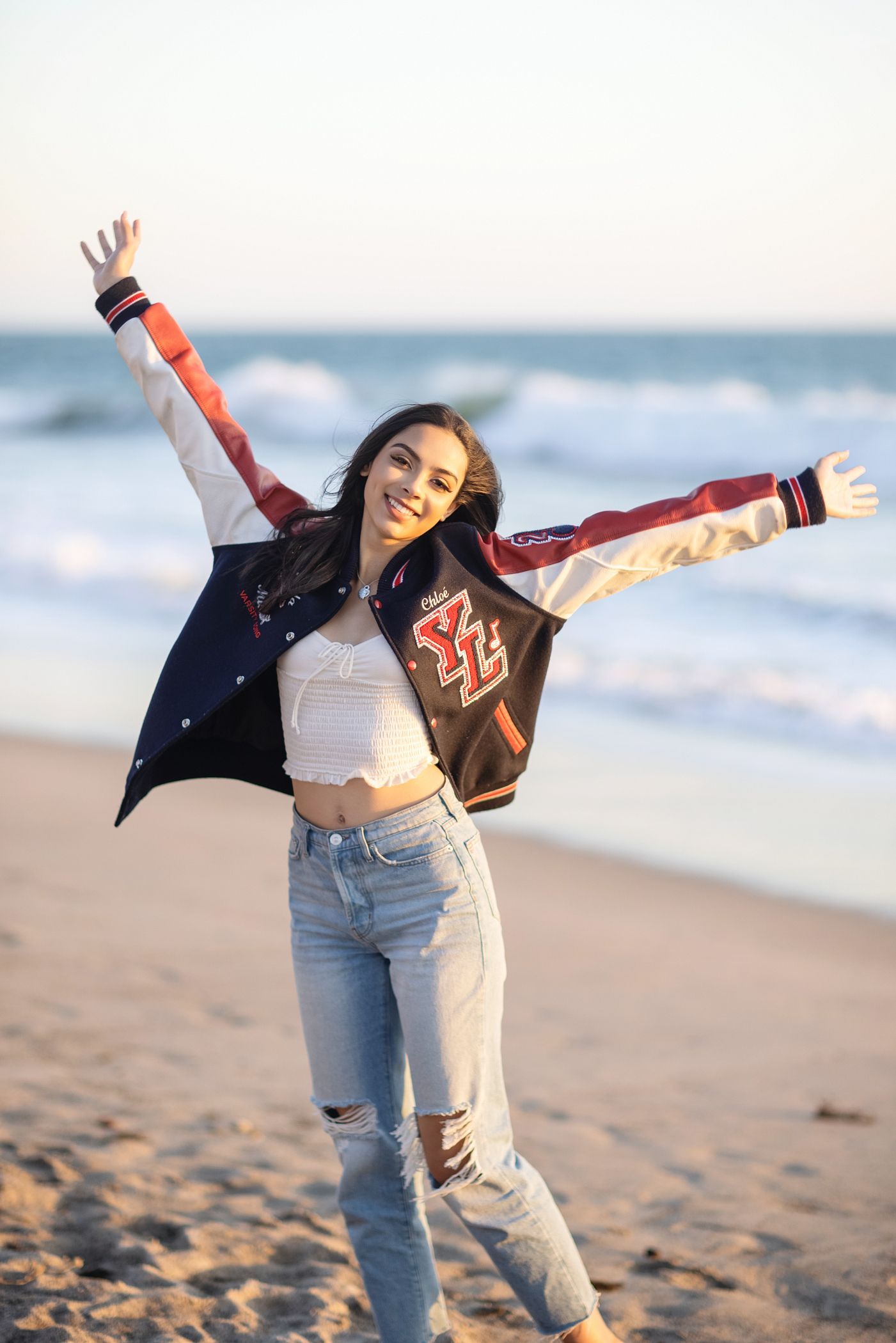 Person with arms raised on a beach, wearing a red jacket and jeans at sunset