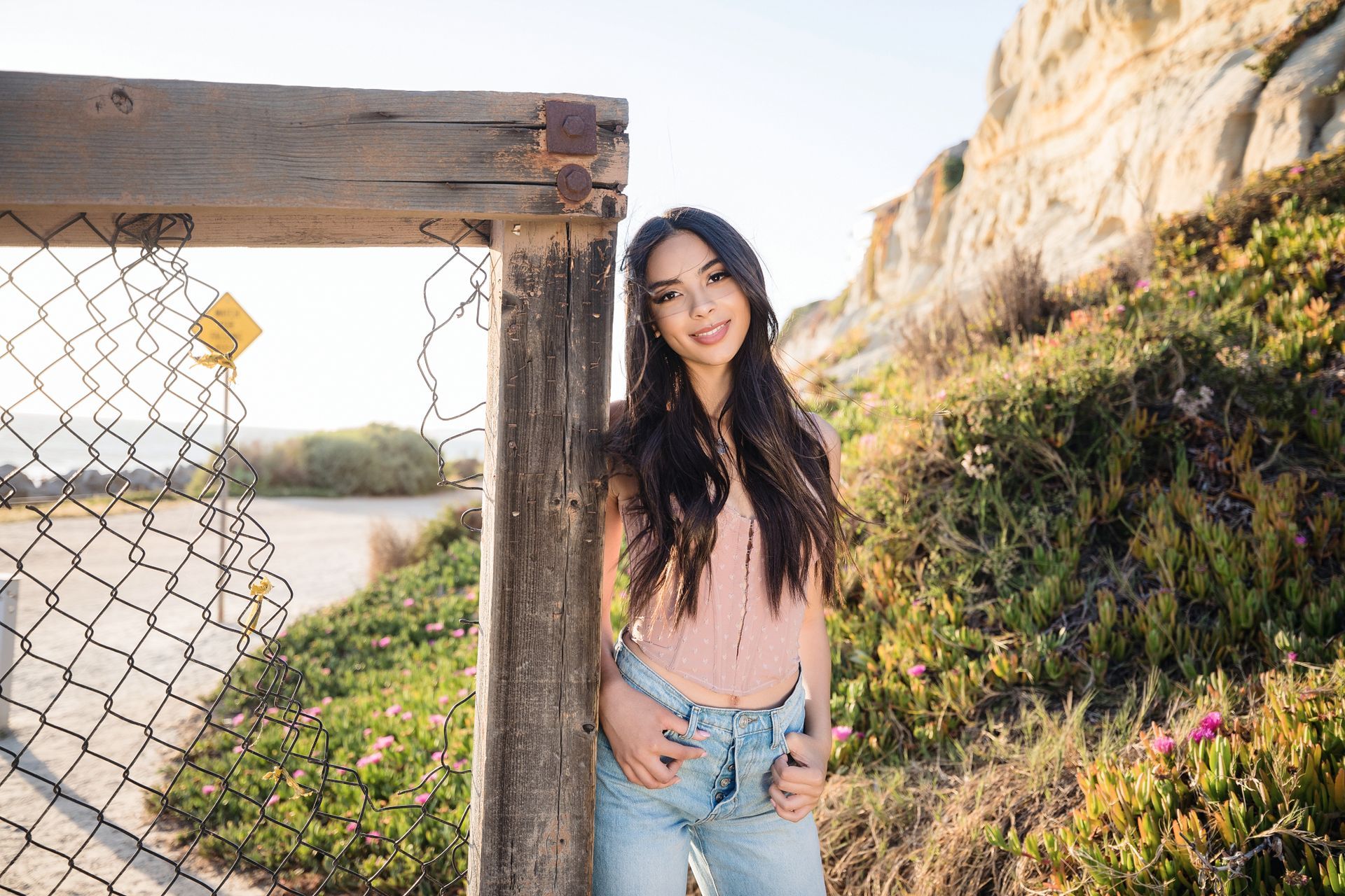 Woman standing by a wooden fence on a sunny coastal path, with cliffs and water in the background.