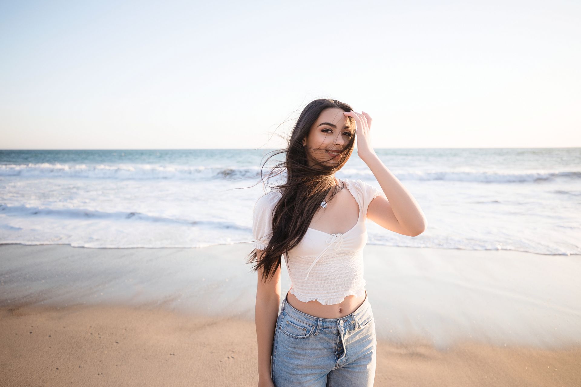 Woman standing on a sandy beach by the ocean, wearing a white top and jeans, with windblown hair.
