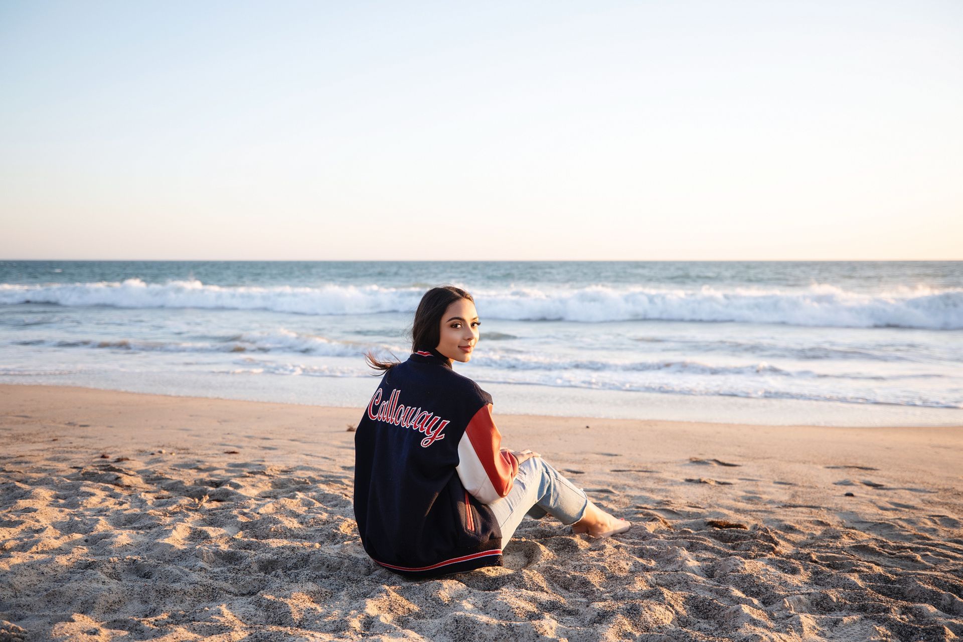 Person sitting on a sandy beach at sunset, facing the ocean with waves in the background