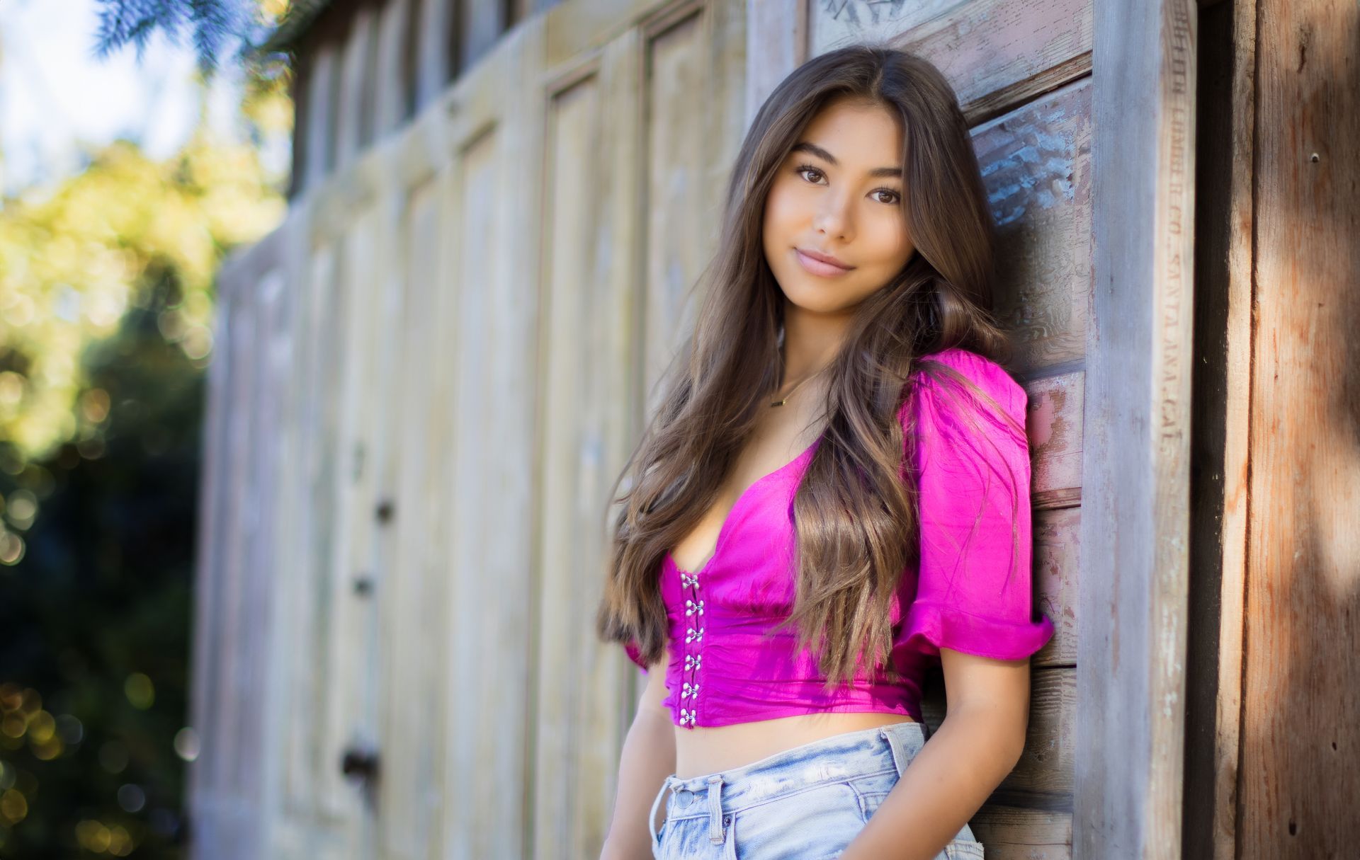 A young woman in a pink crop top and jeans is leaning against a wooden wall.