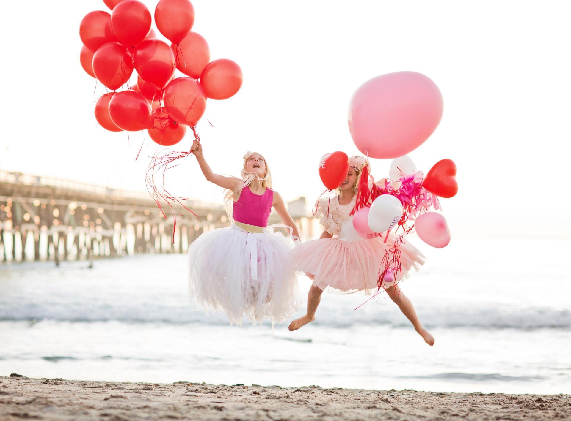 Two girls are jumping in the air while holding balloons on the beach.