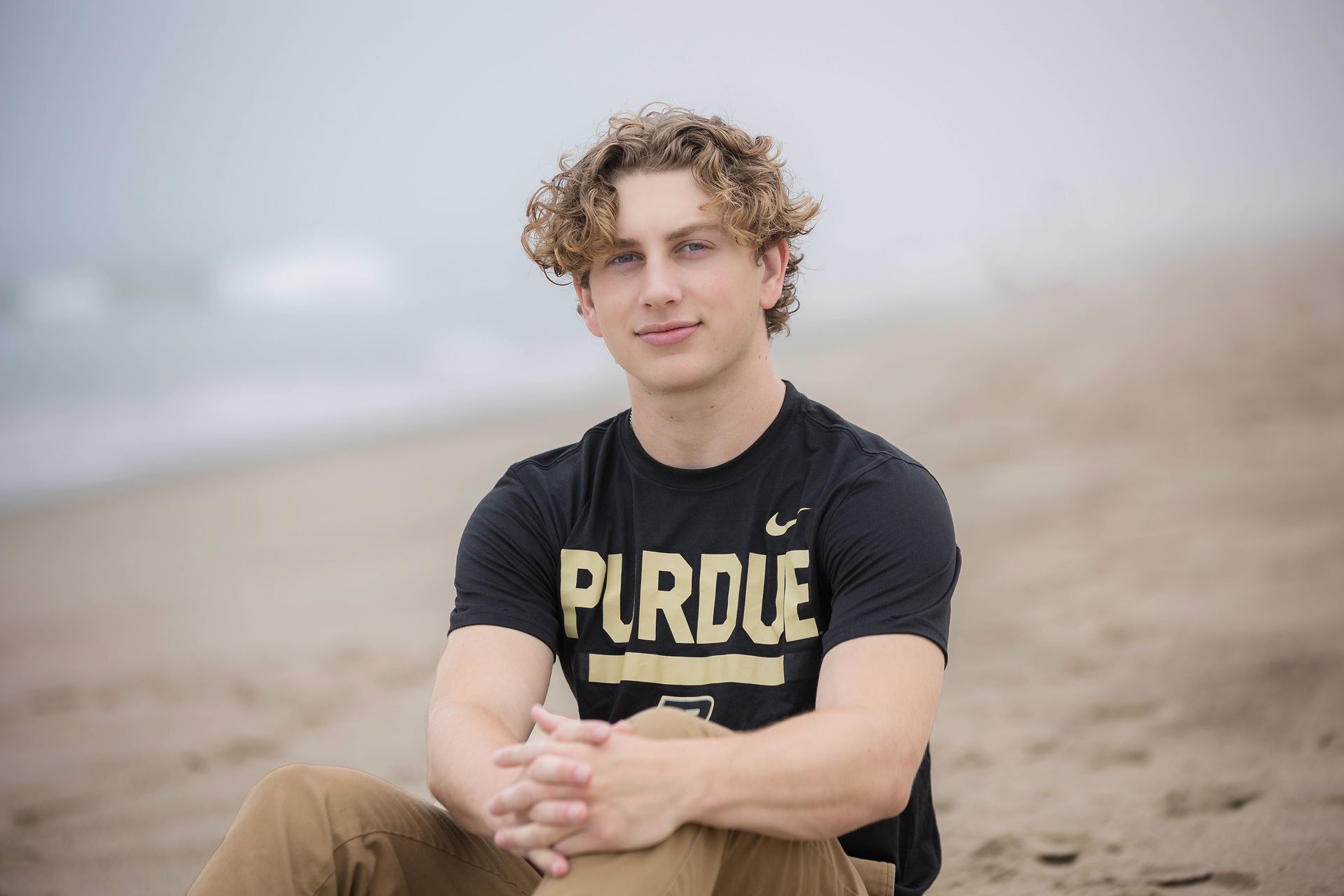 A young man is sitting on the beach wearing a purdue shirt.