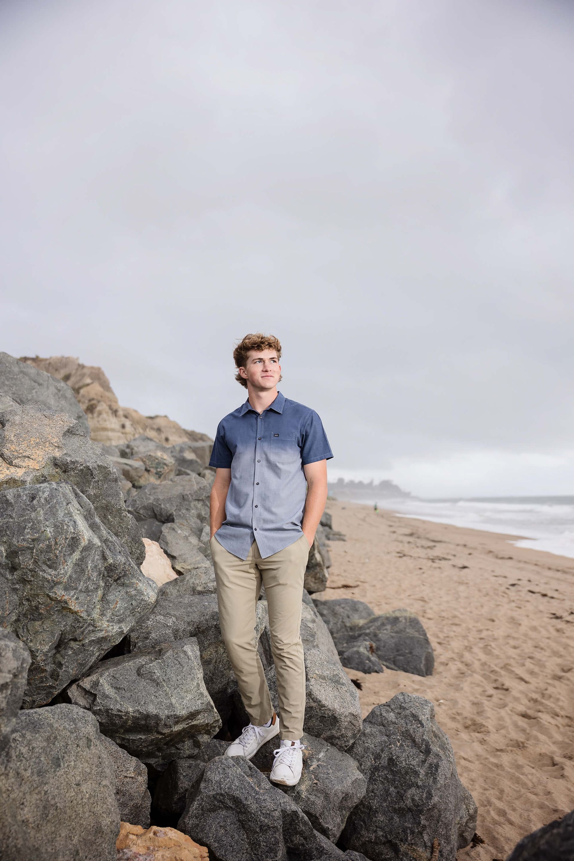 Man standing on seaside rocks beside a sandy beach under a cloudy sky