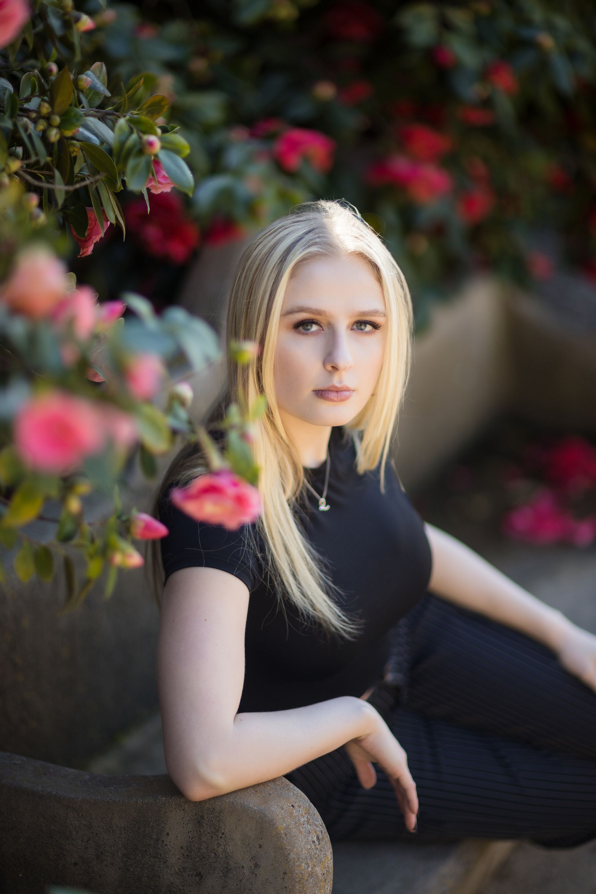 A woman is sitting on a bench in front of a bush of flowers.