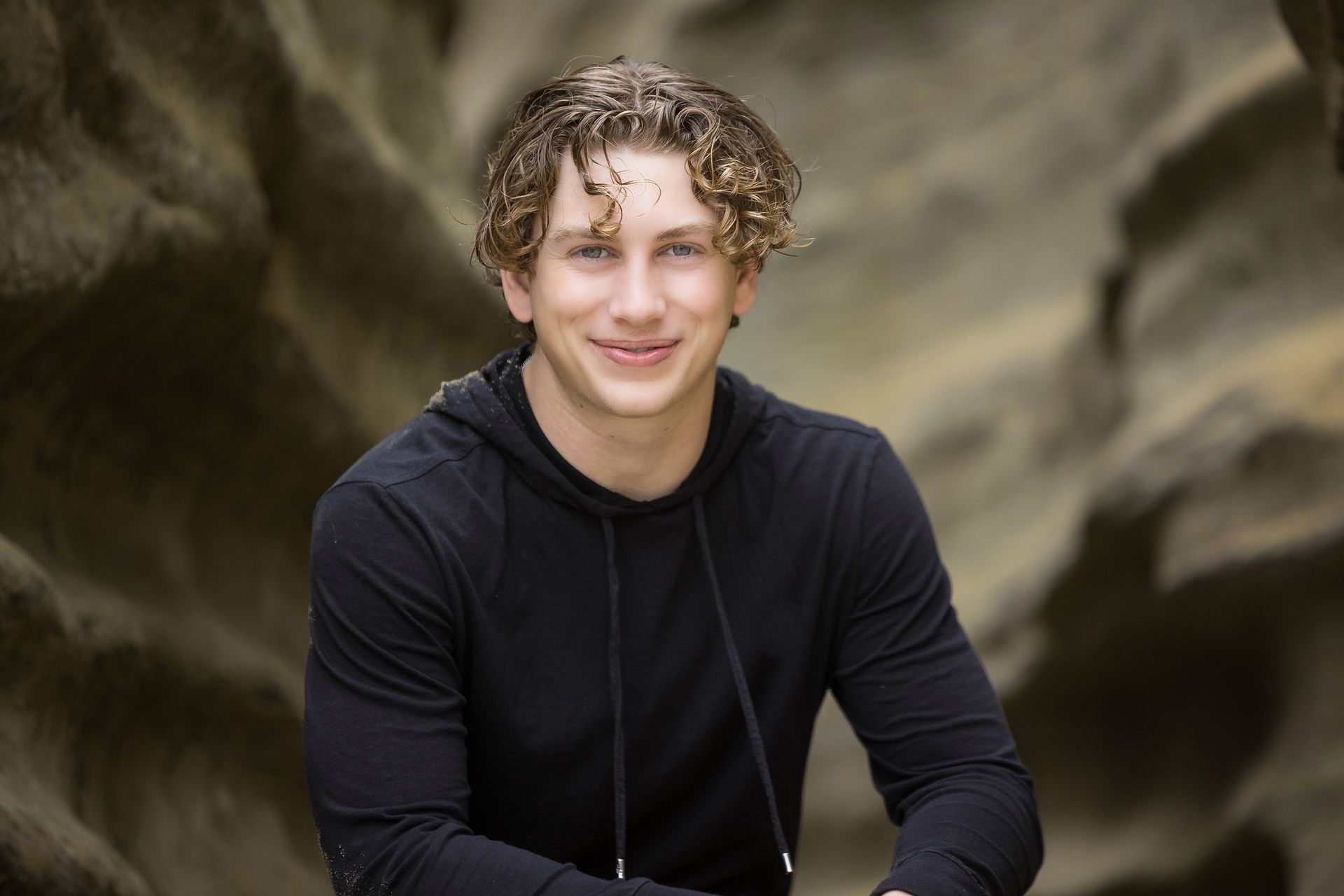 A young man with curly hair is wearing a black hoodie and smiling.