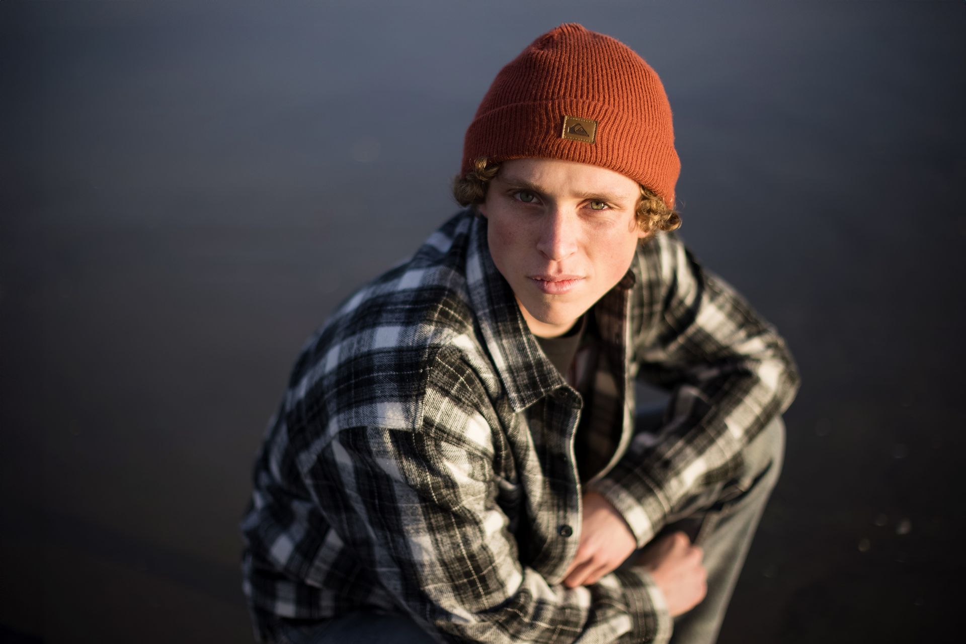 A young man wearing a plaid shirt and a beanie is kneeling down in front of a body of water.