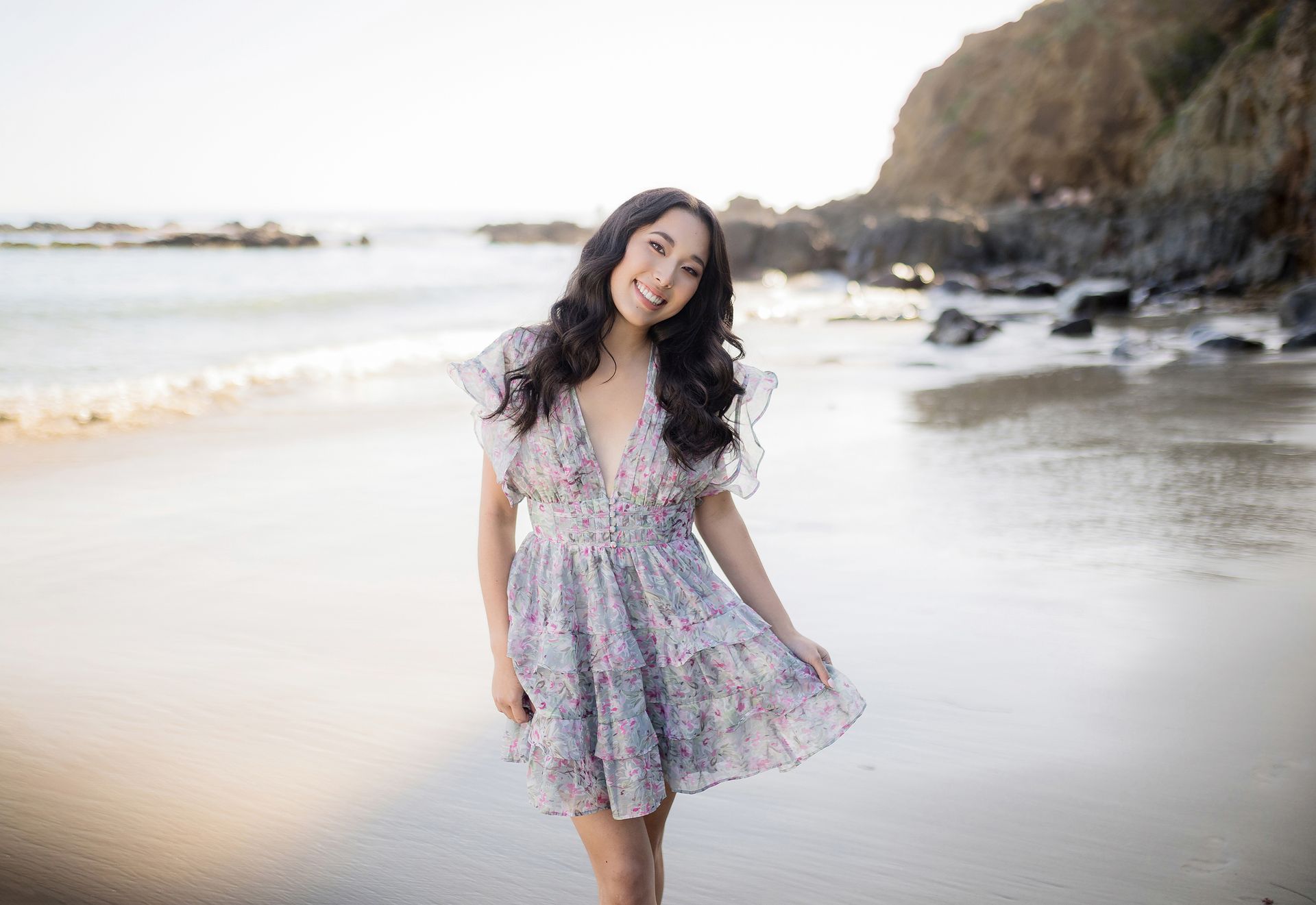 Woman in a floral dress walking on a beach beside rocky cliffs, smiling at the camera.