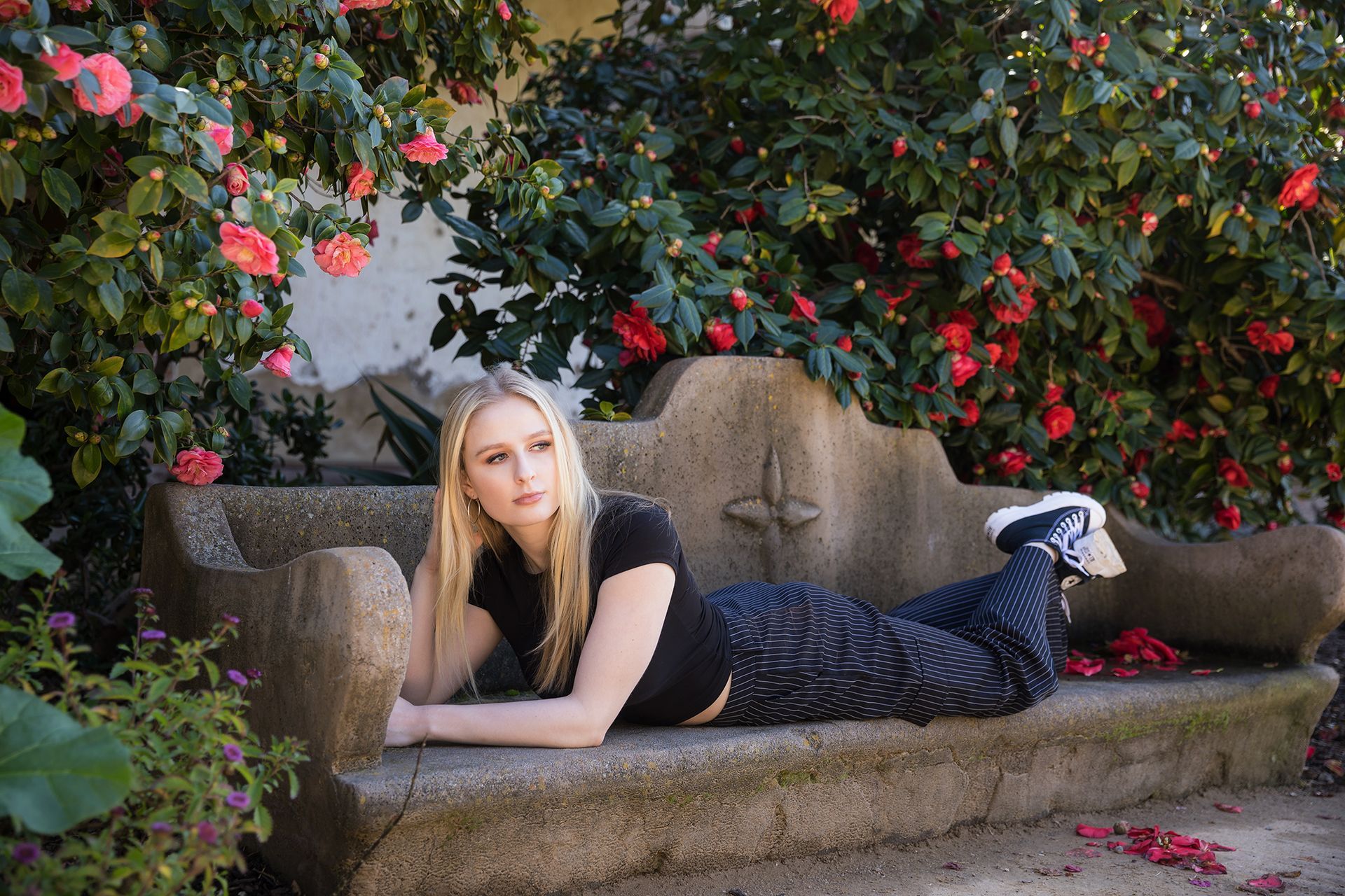A woman is laying on a stone bench in front of a bush of flowers.