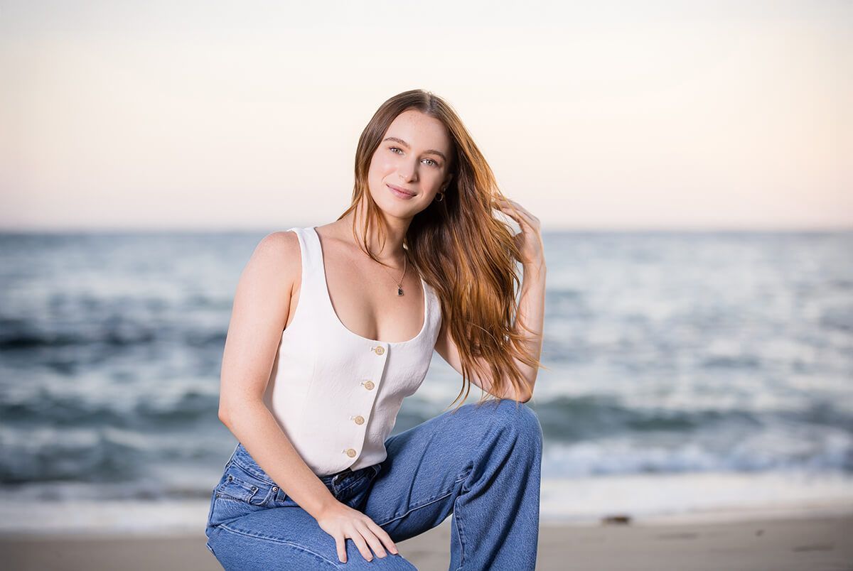 Sunset beach portrait of a young woman with long red hair in jeans and a white tank top,
