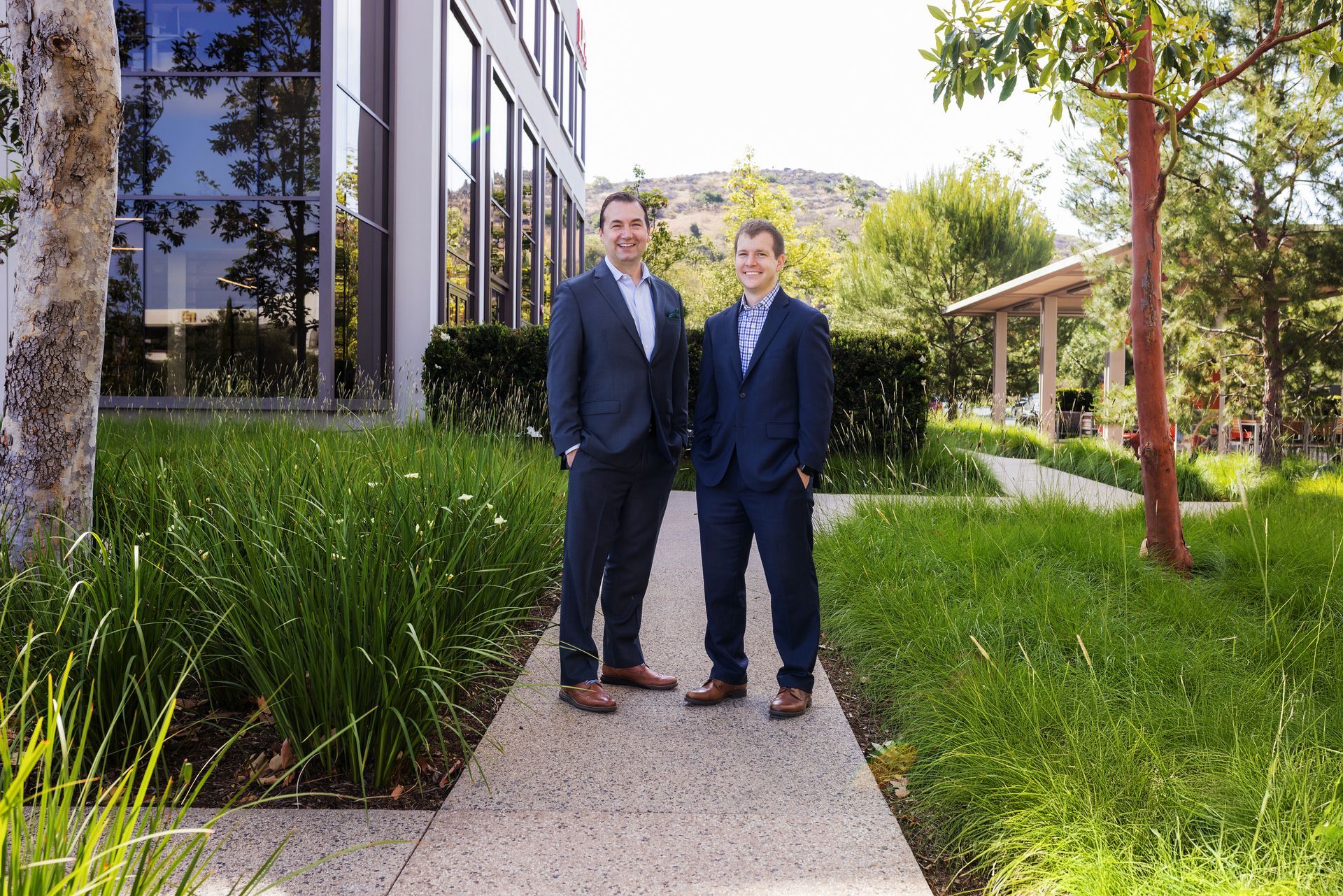 Two men in suits are standing next to each other on a sidewalk in front of a building.