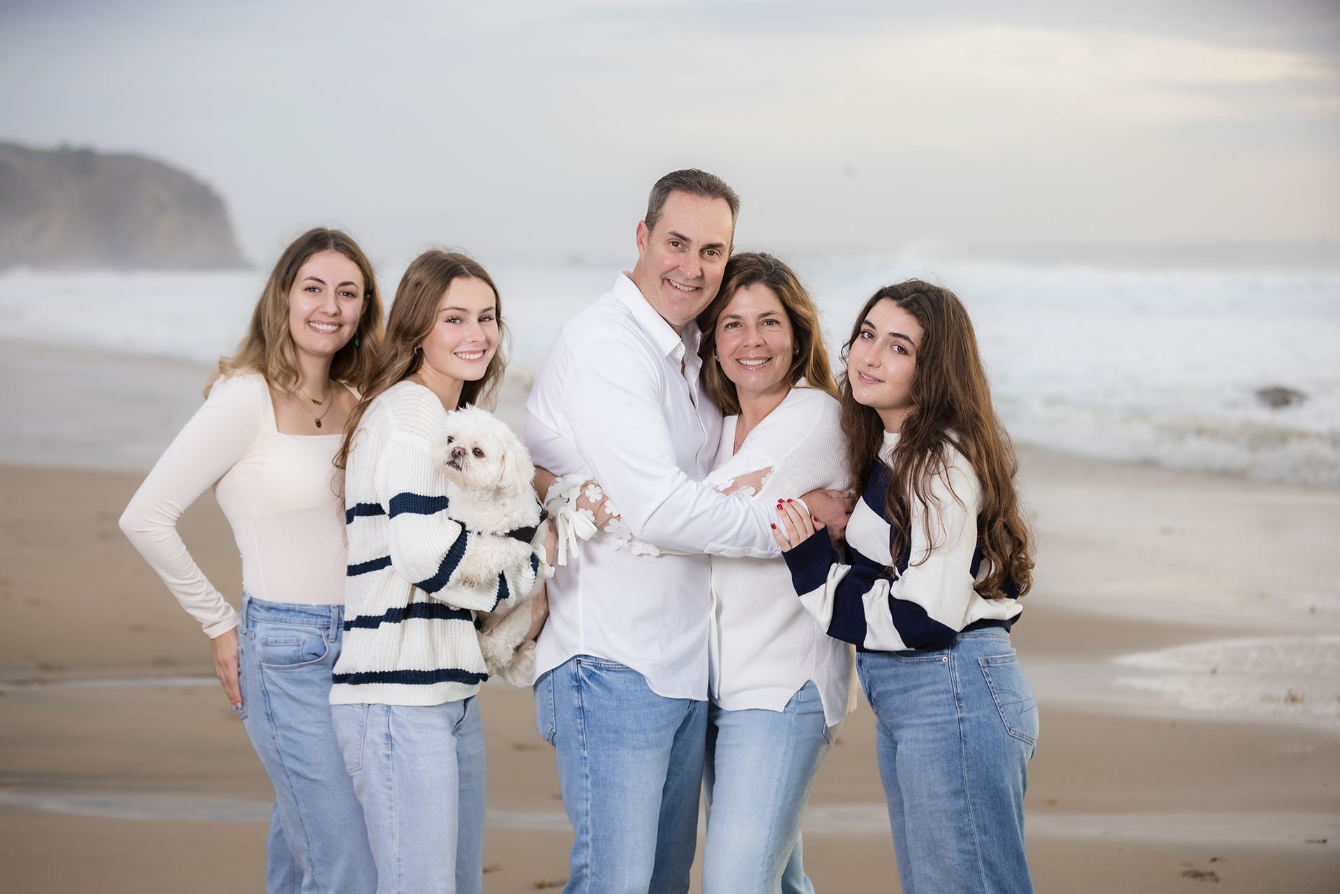 Family smiling on a beach, standing together in white and blue outfits with a small dog