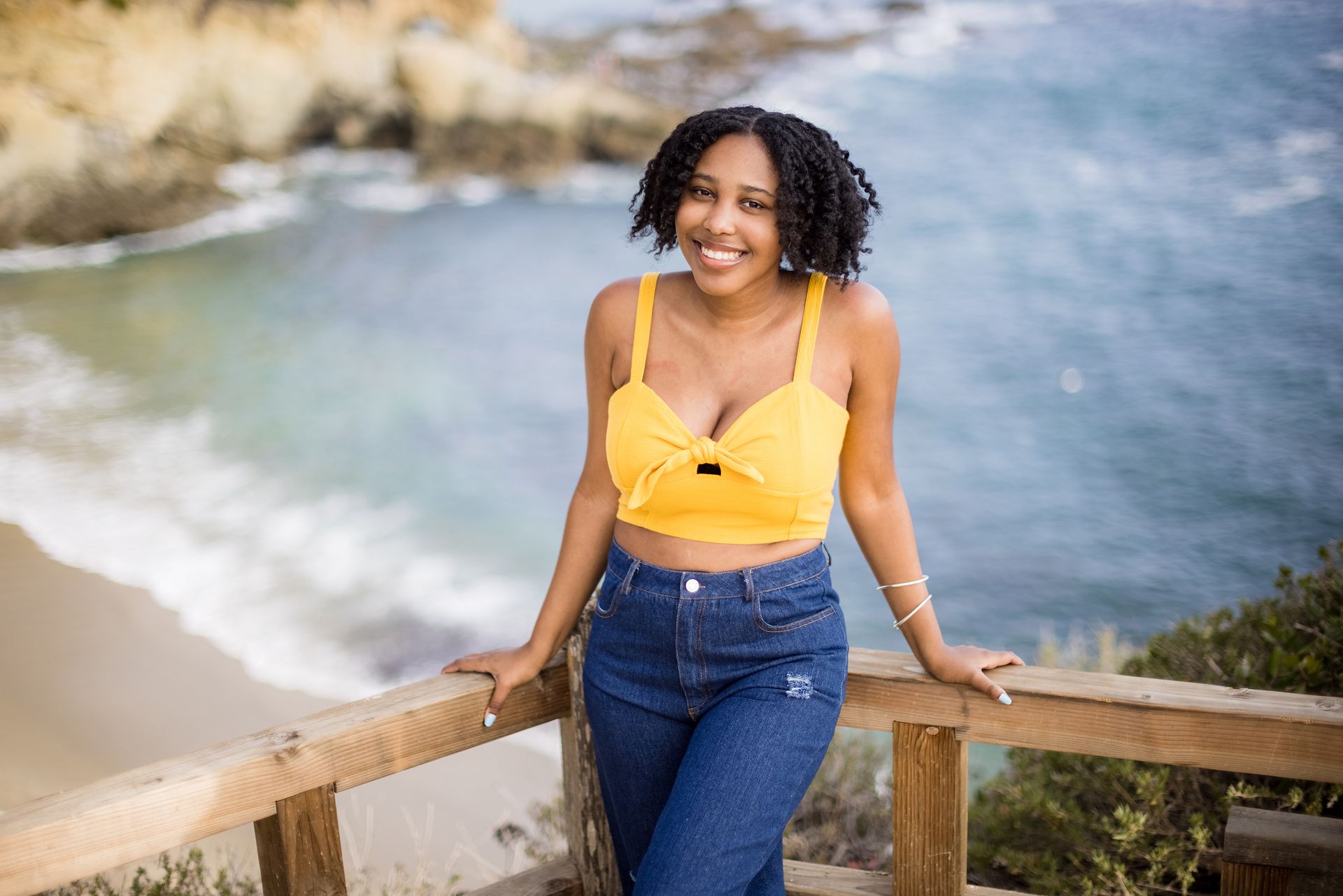 A woman in a yellow top and blue jeans is leaning on a wooden railing overlooking the ocean.