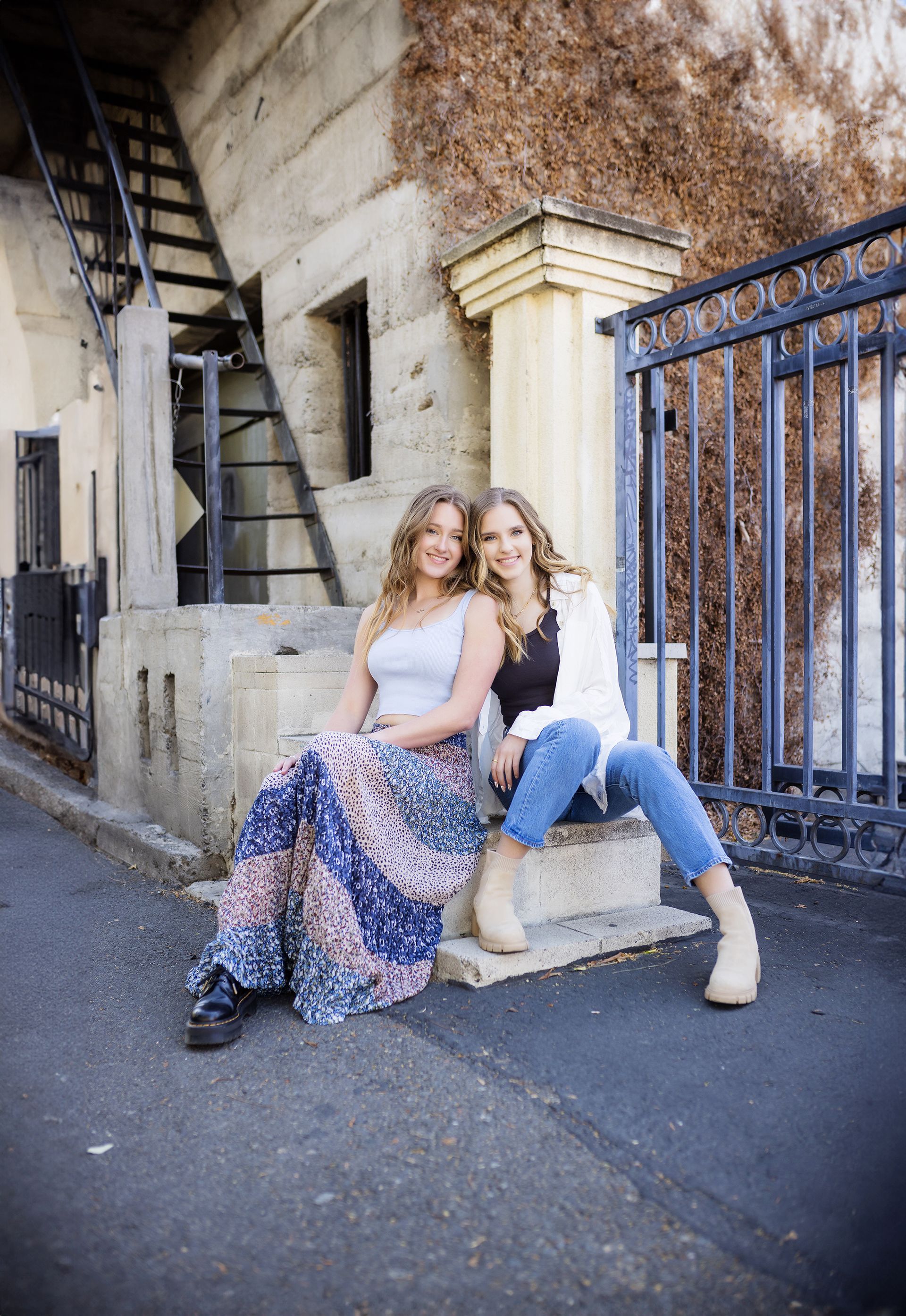 Two women are sitting next to each other on a sidewalk in front of a building.