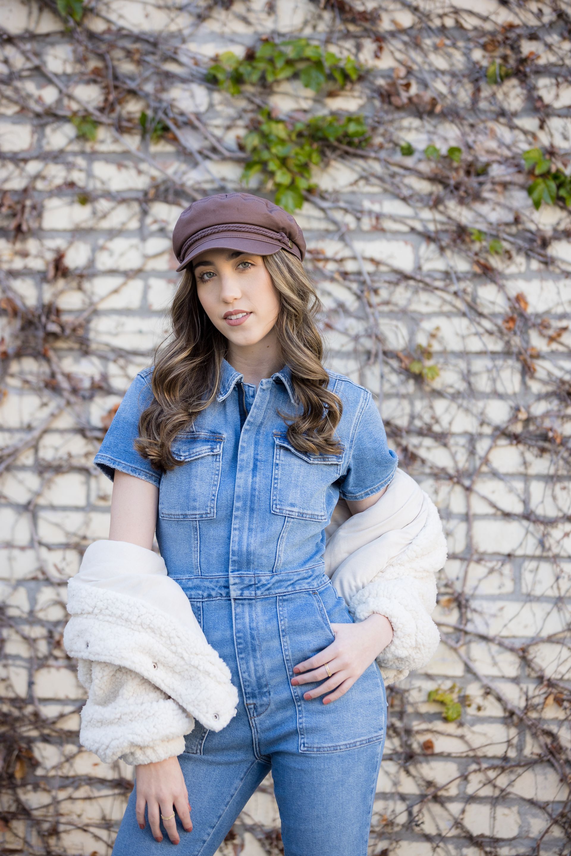 A woman in a denim jumpsuit and hat is standing in front of a brick wall.