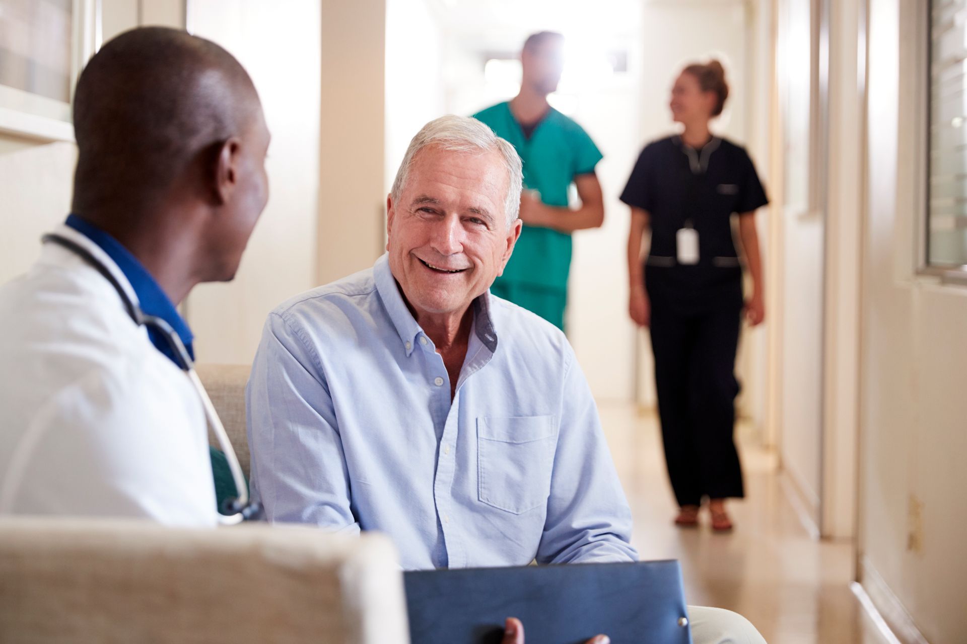 Doctor in white coat converses with smiling older patient; hospital hallway; two healthcare workers in background.