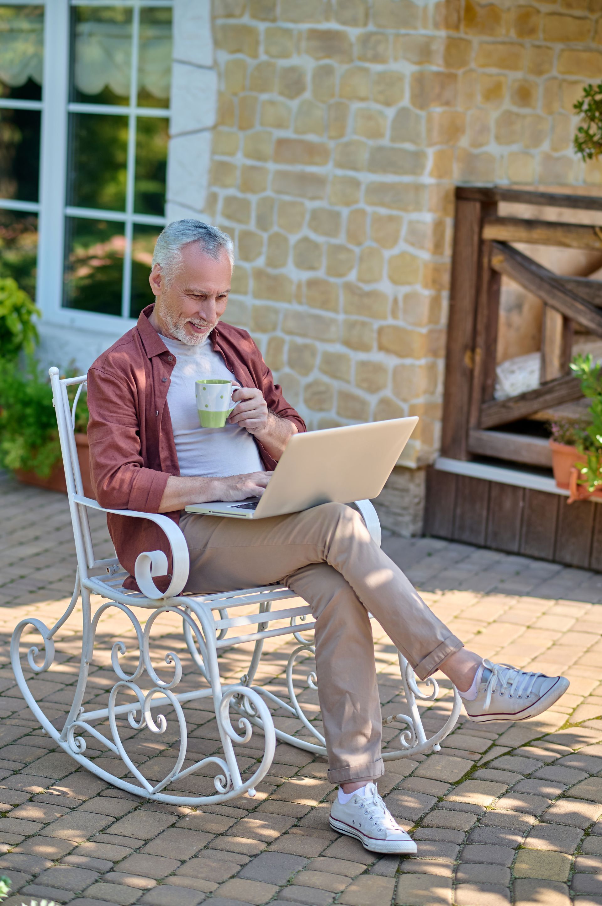 Older man, gray hair, using laptop, drinking coffee, sitting on white rocking chair outdoors.