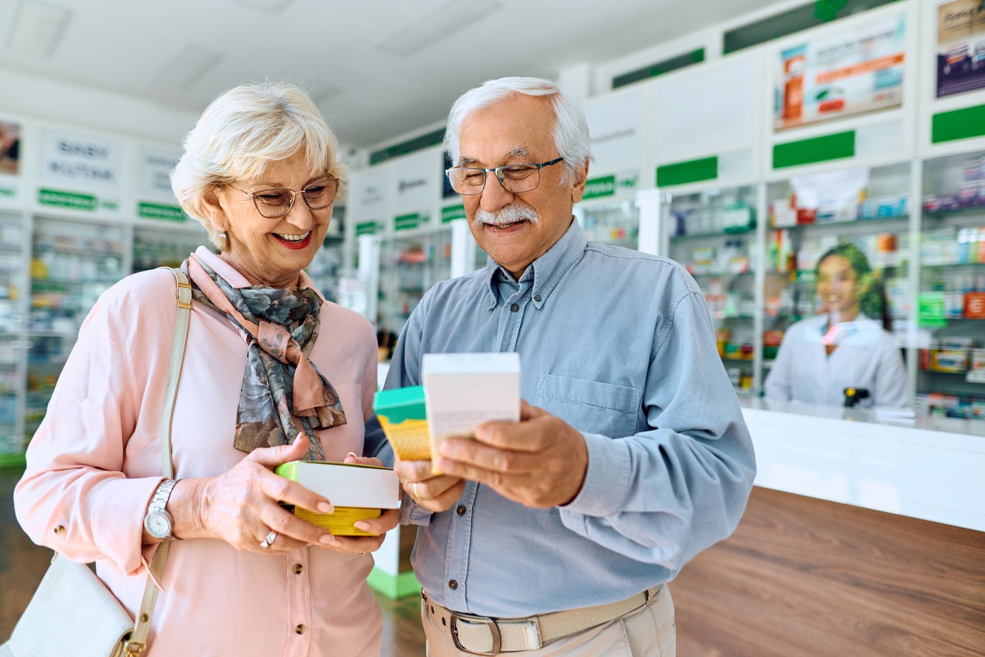 Senior couple in pharmacy examining medication boxes; pharmacist in background.