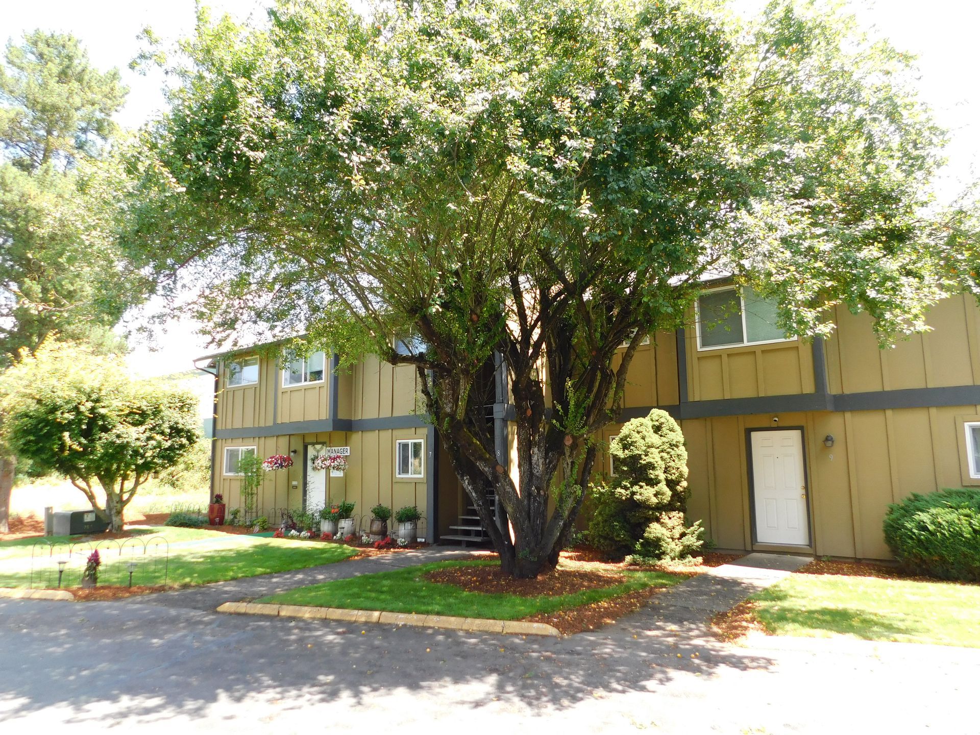 Two-story brown building with green trees and shrubs in front. Sunny day.