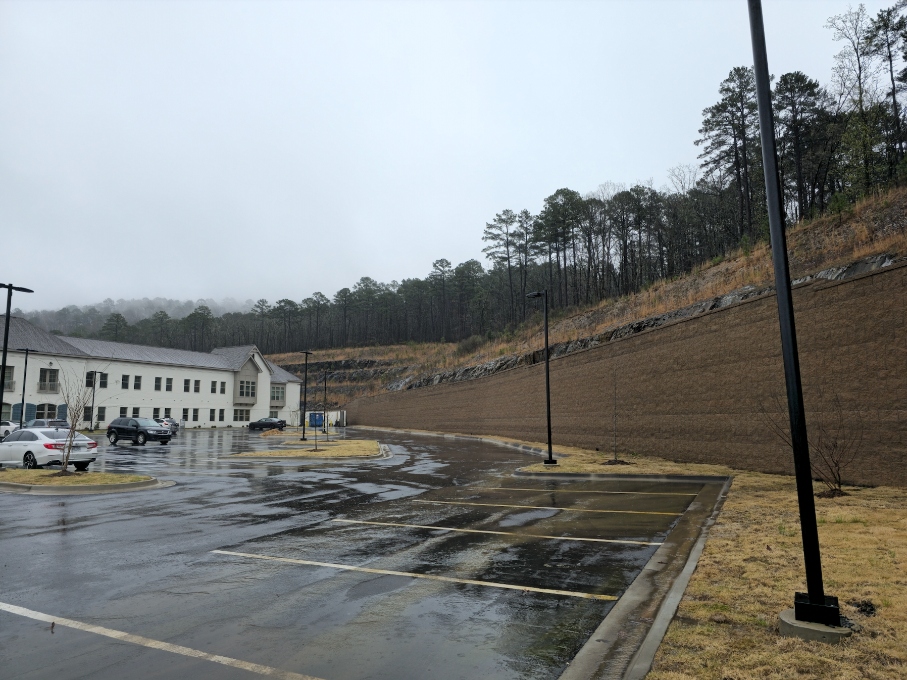 Rainy parking lot beside white buildings and a steep wooded hillside, with a retaining wall and lampposts.