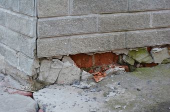 Cracked brick foundation with crumbling concrete and exposed red bricks near the corner of a wall