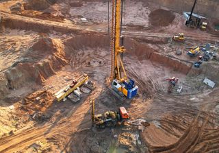 Excavator and drilling rig working in a muddy construction pit with workers and equipment nearby.