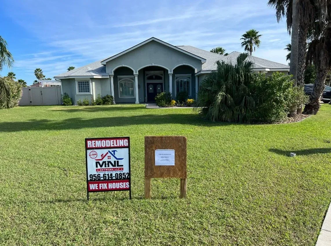 House with two yard signs on a sunny lawn, one partially obscured by a bush.