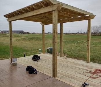 Wooden pergola on a deck in a grassy field, with tools and cords scattered on the floor.