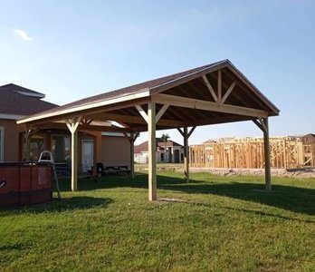 Wooden backyard carport with a gabled roof on a grassy lawn, with a house and fence in the background