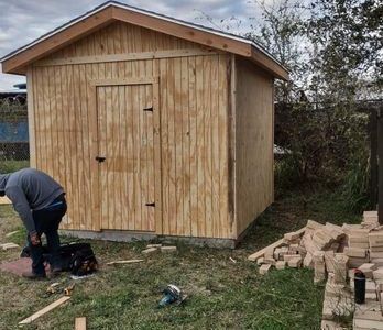 Person bending near a wooden shed while stacked bricks and tools sit on the grass nearby.