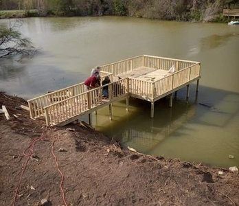 Wooden dock under construction over a muddy pond, with a person working on it from the shore.