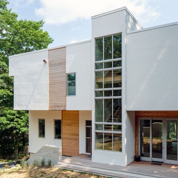 Modern white house with large glass windows and a wood-accented entrance amid green trees