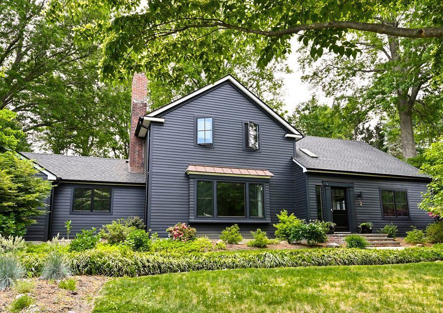 Dark gray house with white trim and brick chimney, surrounded by lush green trees and garden landscaping