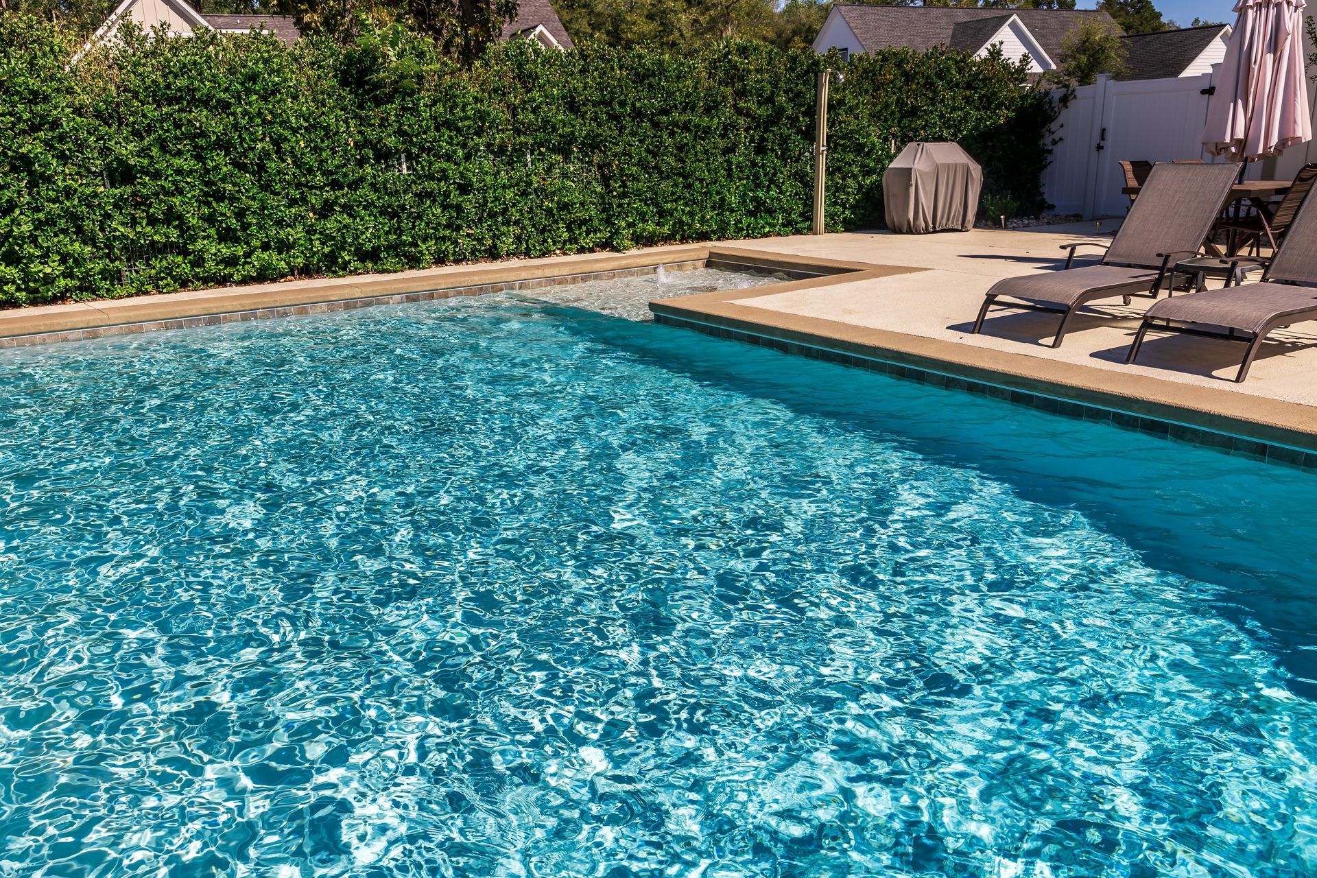 Outdoor swimming pool with sparkling blue water, lounge chairs, and patio umbrellas beside a hedge-lined deck