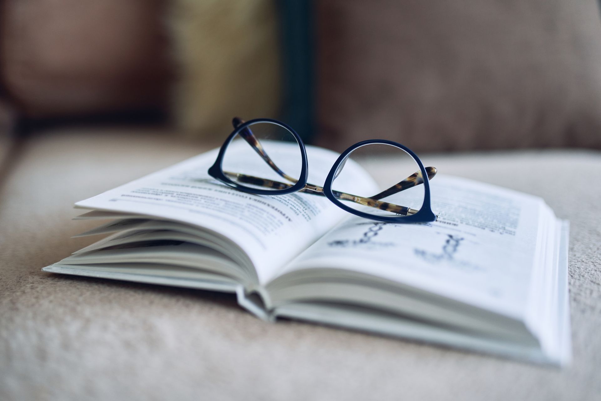 Open book with eyeglasses resting on the pages, indoors on a sofa.