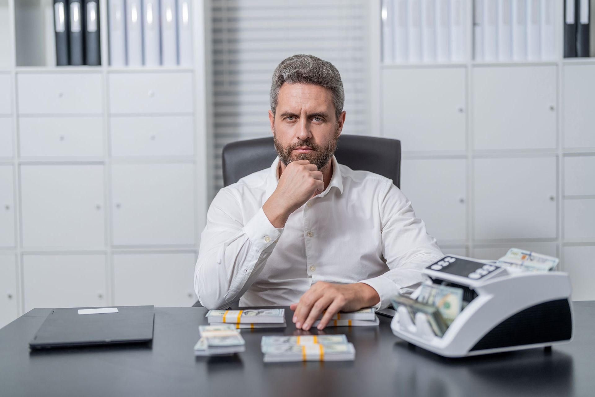 A man in white shirt at desk, hand on chin, beside stacked cash bundles and a bill-counting machine. A man in white shirt at desk, hand on chin, beside stacked cash bundles and a bill-counting machine.
