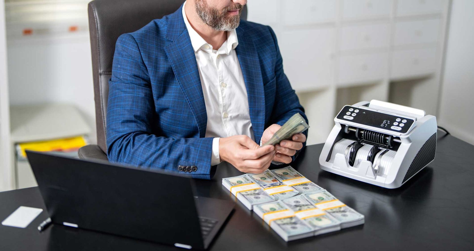 A man in blazer counting cash beside a bill-counting machine with banknote stacks on an office desk. A man in blazer counting cash beside a bill-counting machine with banknote stacks on an office desk.