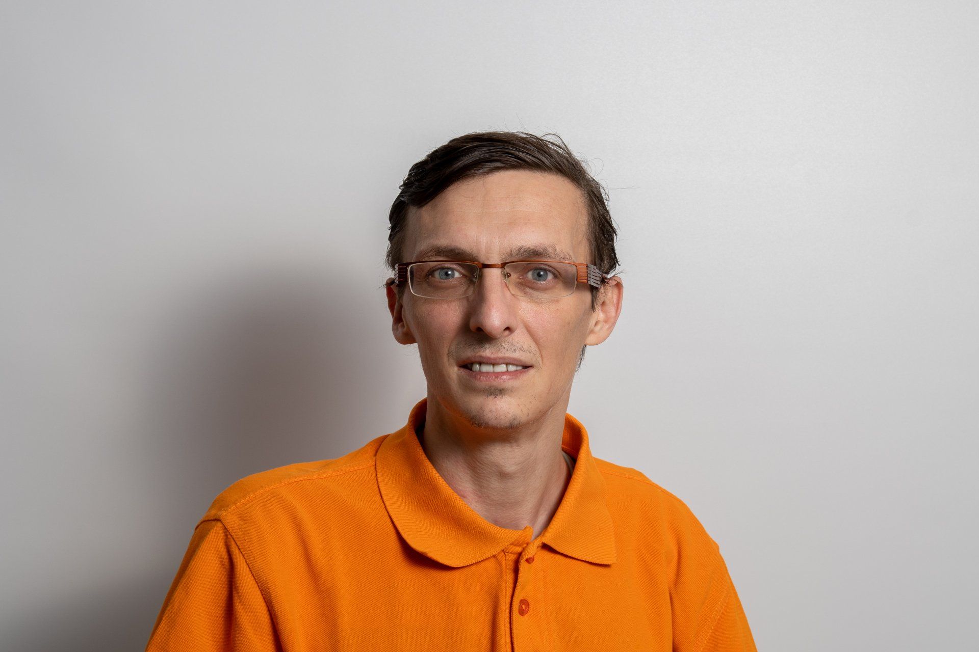 Ein Mann mit Brille und orangefarbenem Poloshirt steht vor einer weißen Wand.