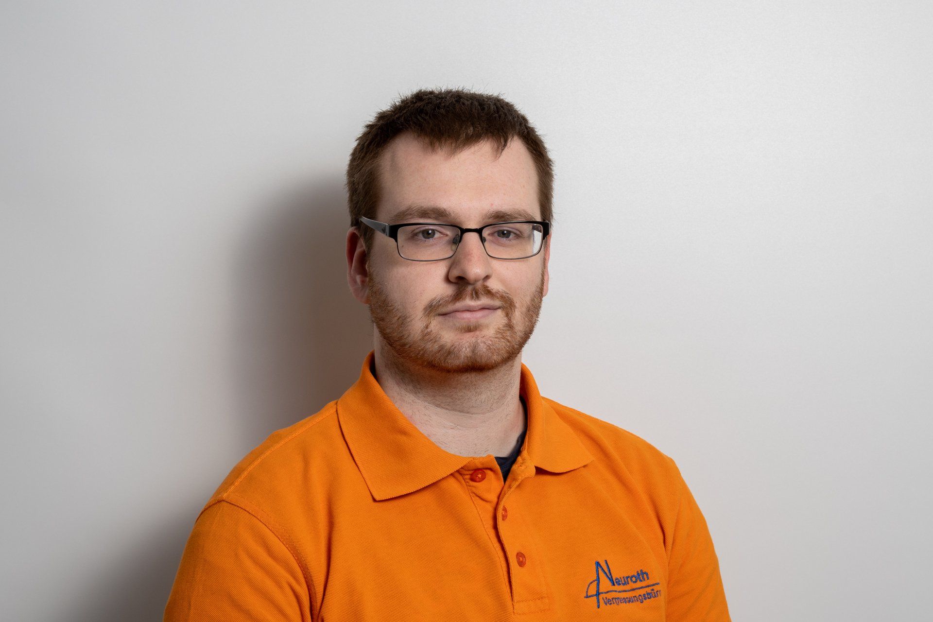 Ein Mann mit Brille und orangefarbenem Poloshirt steht vor einer weißen Wand.