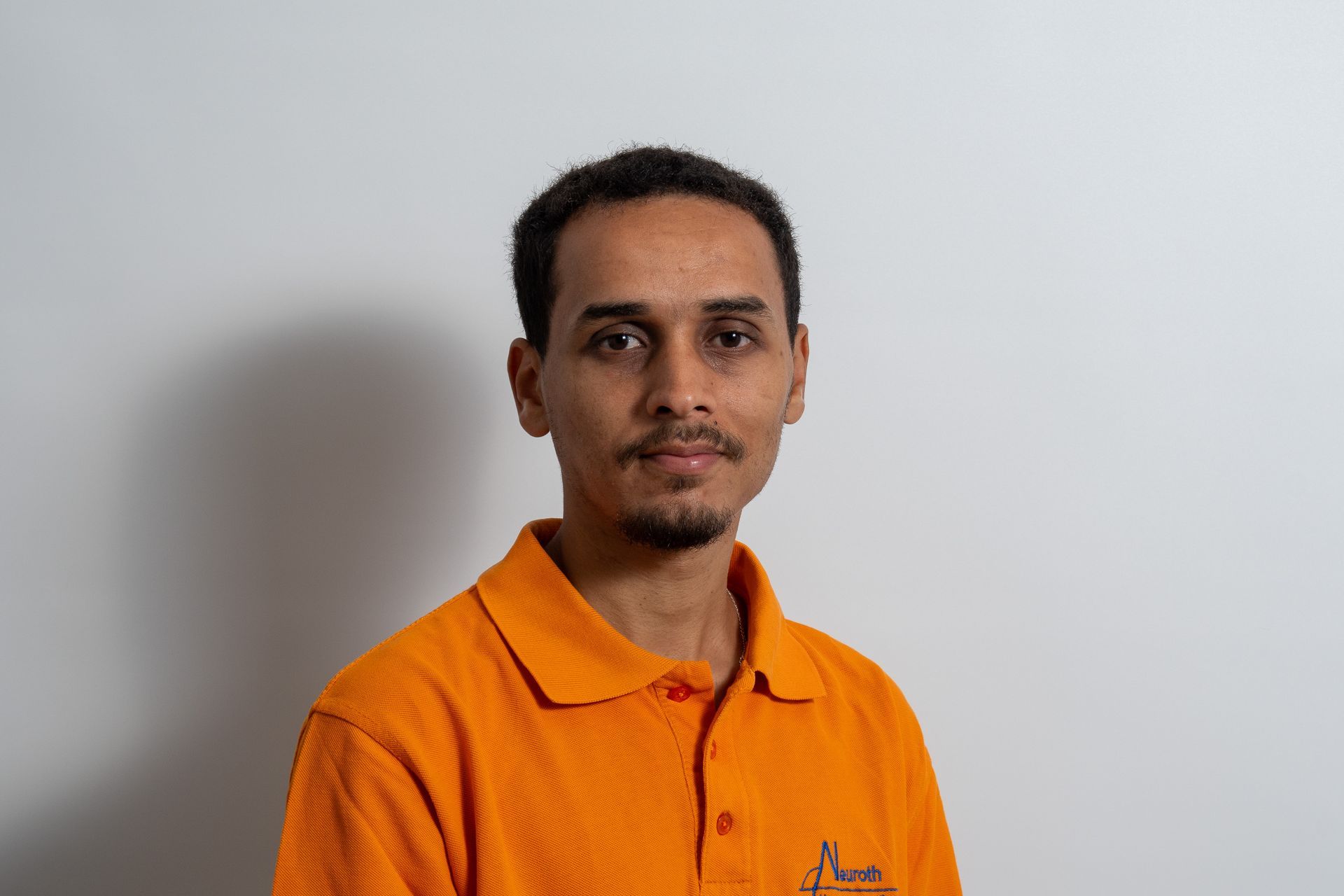 Ein Mann in einem orangefarbenen Poloshirt steht vor einer weißen Wand.