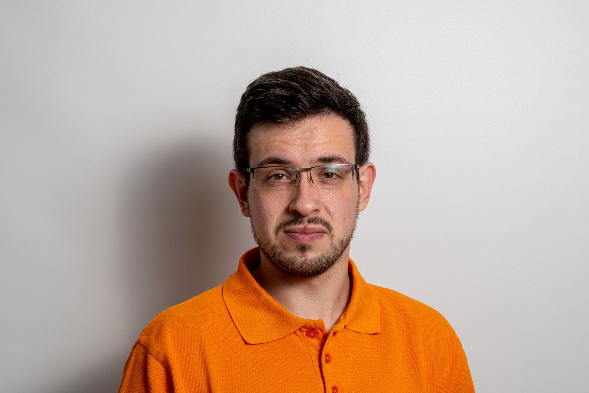 Ein Mann mit Brille und orangefarbenem Poloshirt steht vor einer weißen Wand.