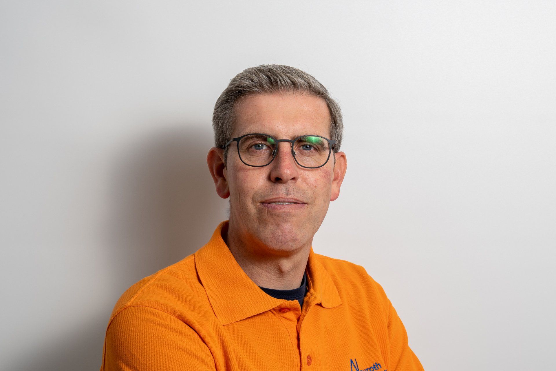 Ein Mann mit Brille und orangefarbenem Hemd steht vor einer weißen Wand.