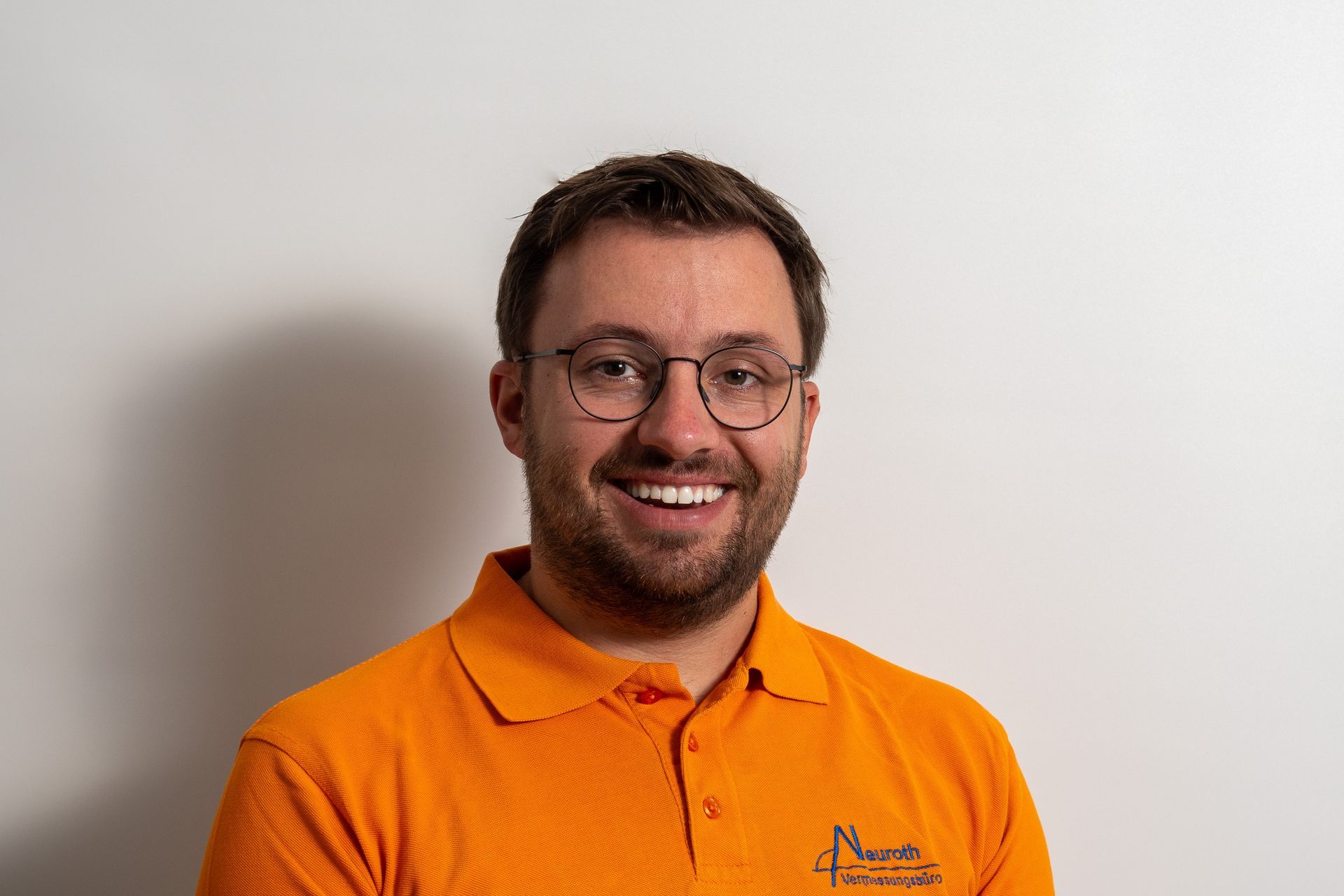 Ein Mann mit Brille und orangefarbenem Poloshirt lächelt in die Kamera.