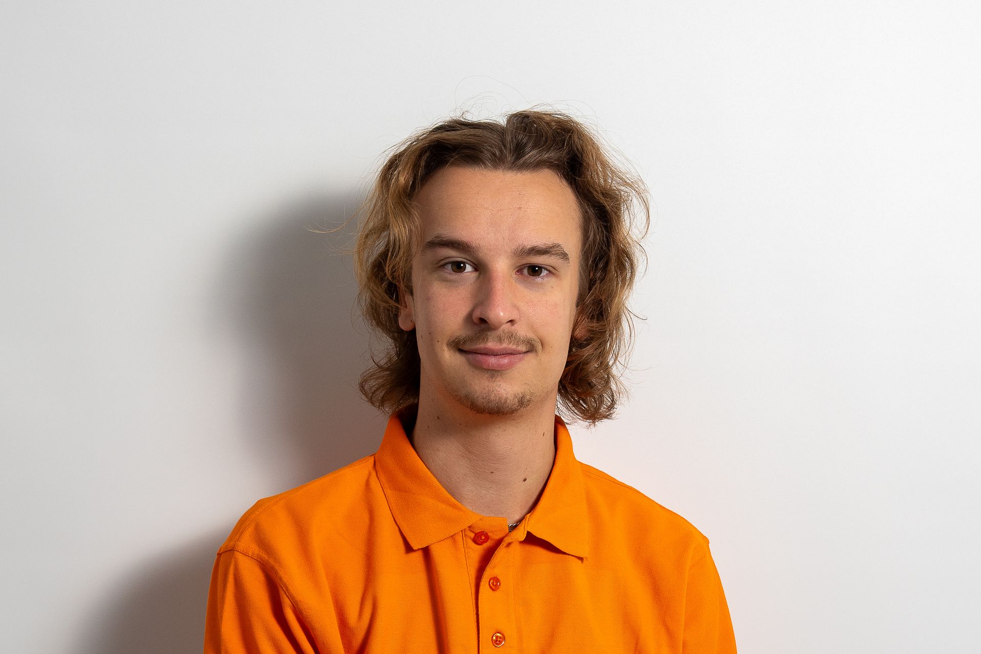 Ein junger Mann mit lockigem Haar trägt ein orangefarbenes Poloshirt.
