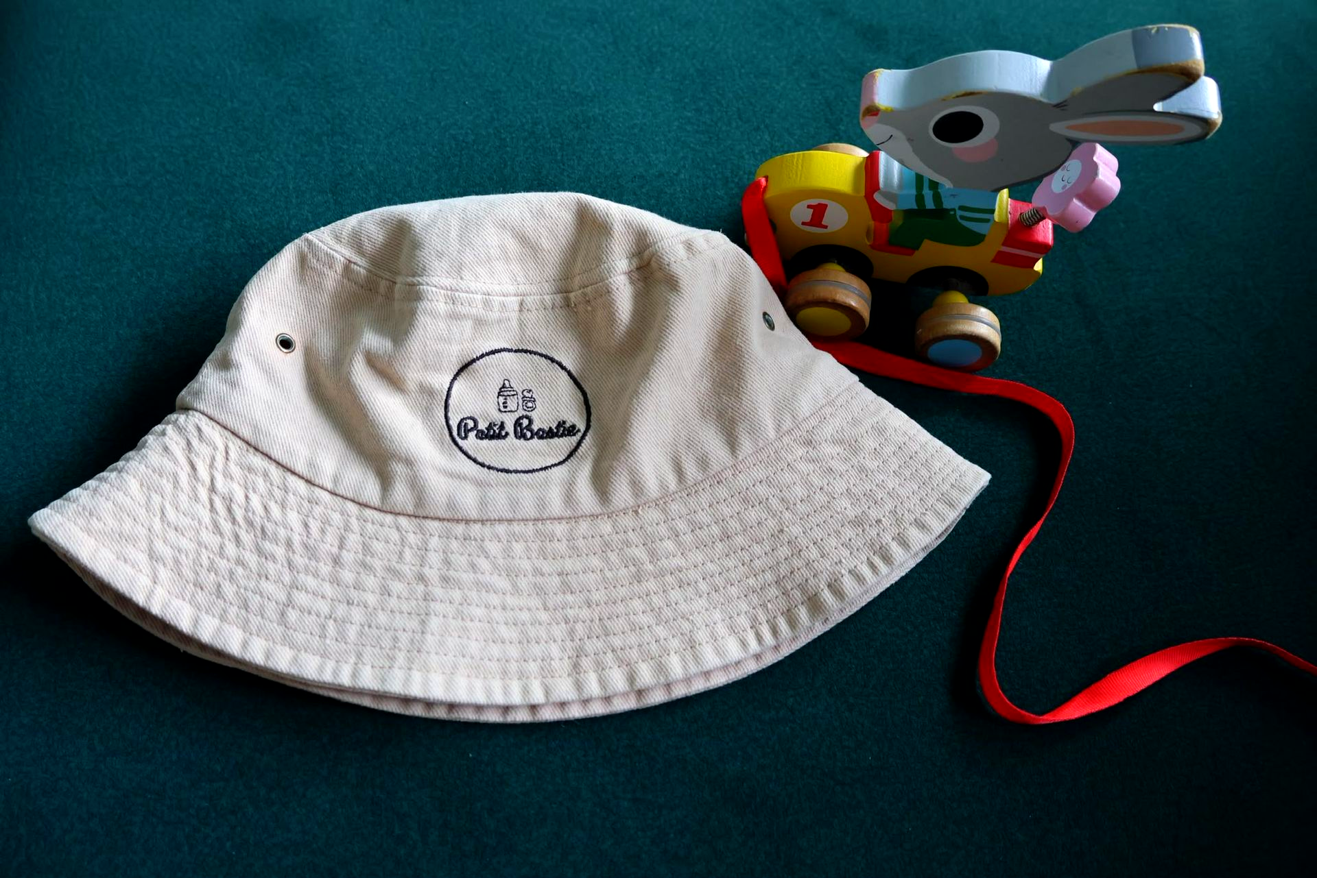 Beige bucket hat with a round logo beside a small toy on a teal surface, with a red string attached