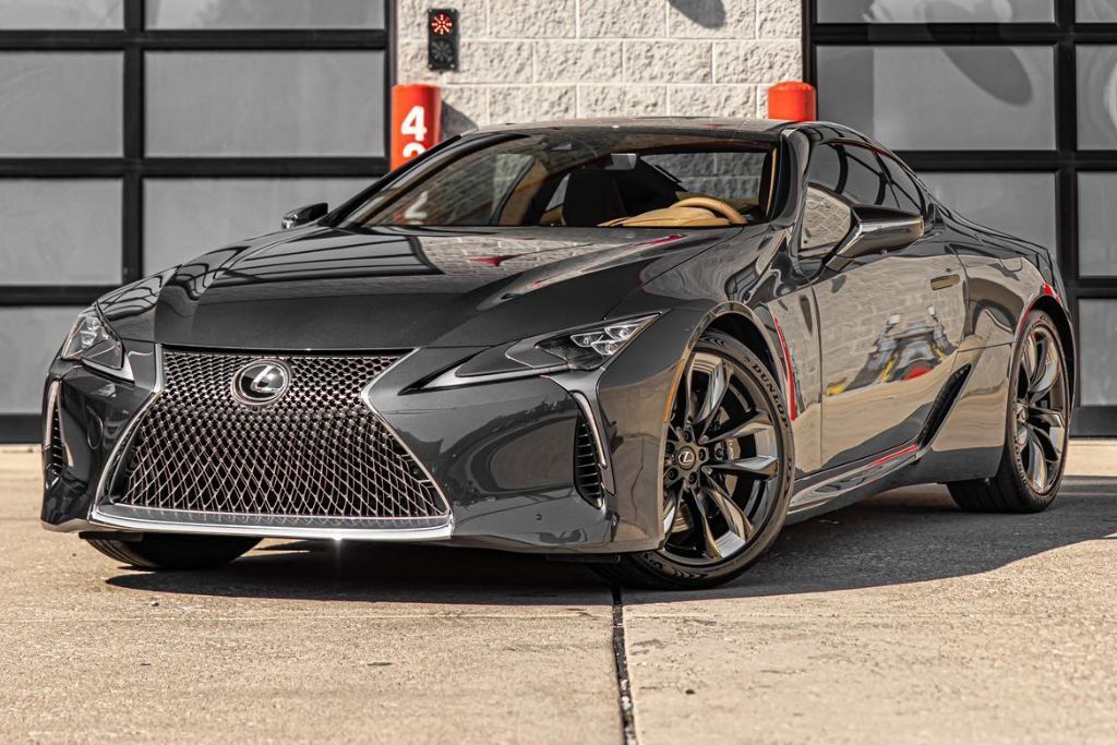 image displays a sleek, black Lexus coupe parked in front of a garage with glass-paneled doors. The vehicle is highlighted by its bold, spindle-shaped front grille, sculpted body lines, and distinctive chrome accents. The sporty and luxurious design of the car is enhanced by its aggressive stance and stylish alloy wheels.