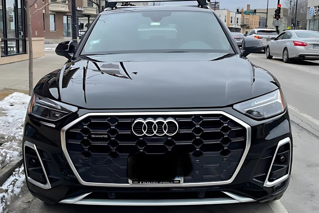 image shows the front view of a black Audi SUV parked on a city street, showcasing its bold honeycomb grille with the Audi logo prominently displayed. The vehicle’s glossy finish reflects the surrounding buildings and street, suggesting a recently applied protective coating. Snow can be seen on the sidewalk, contrasting with the sleek appearance of the car.