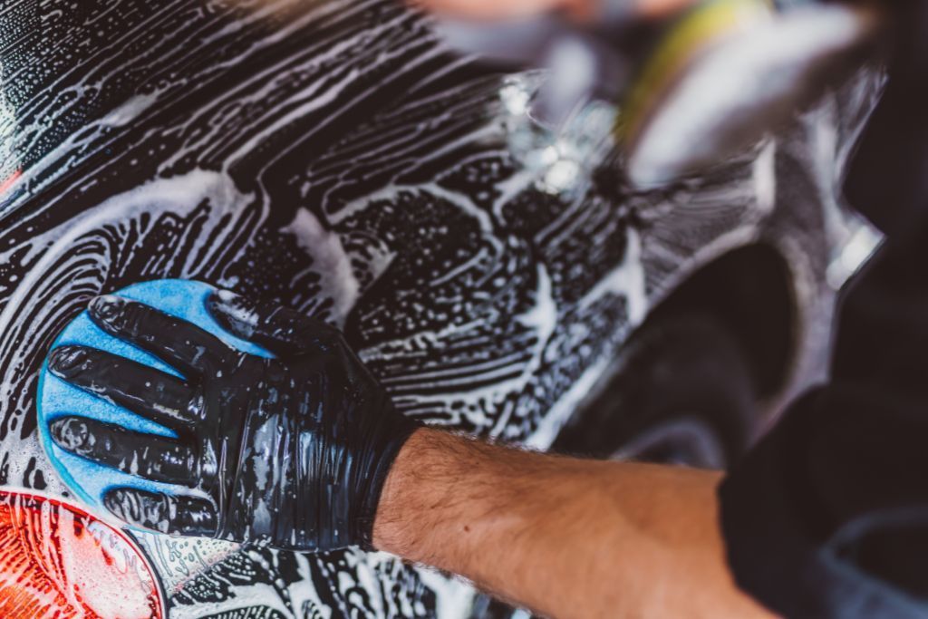 A person wearing black gloves uses a blue sponge to wash a car's surface, covering it with soap suds during the preparation for ceramic coating.