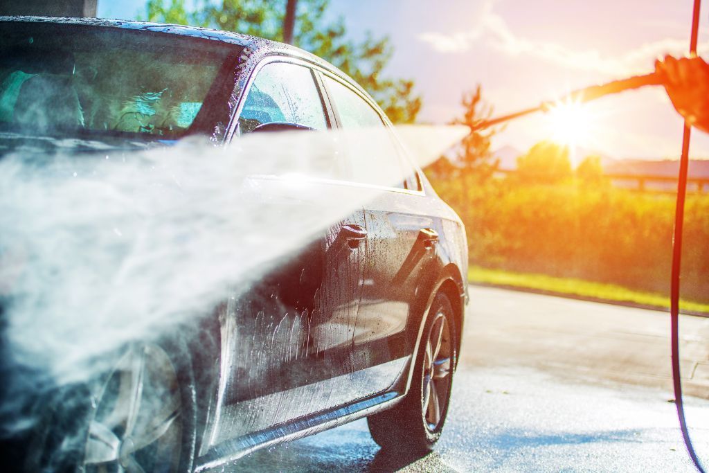 A car being washed with a pressure hose under bright sunlight, highlighting the ease of maintenance provided by ceramic coating. The water streams off the vehicle, emphasizing the hydrophobic properties that reduce dirt buildup and washing frequency.