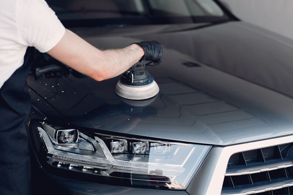 A person wearing a white shirt and black gloves is using a power buffer to polish the hood of a dark-colored car, focusing on achieving a smooth, reflective finish on the vehicle's surface.